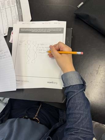 A photo taken over the shoulder of a person seated at a black table, using their right hand to draw on a printed worksheet. Loose papers and a black fabric pouch sit near the top-right edge of the photo. A photo taken over the shoulder of a person seated at a black table, using their right hand to draw on a printed worksheet. Loose papers and a black fabric pouch sit near the top-right edge of the photo.