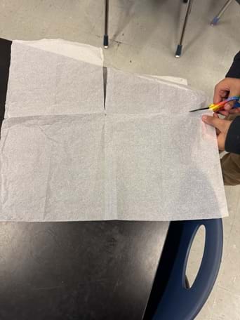 A photo showing an overhead view of a student cutting a large sheet of thin white tissue paper on a black tabletop. Two hands hold rainbow-handled scissors at the right edge of the sheet; a straight cut has been made near the top center. The scene suggests students building paper strap prototypes. A photo showing an overhead view of a student cutting a large sheet of thin white tissue paper on a black tabletop. Two hands hold rainbow-handled scissors at the right edge of the sheet; a straight cut has been made near the top center. The scene suggests students building paper strap prototypes.