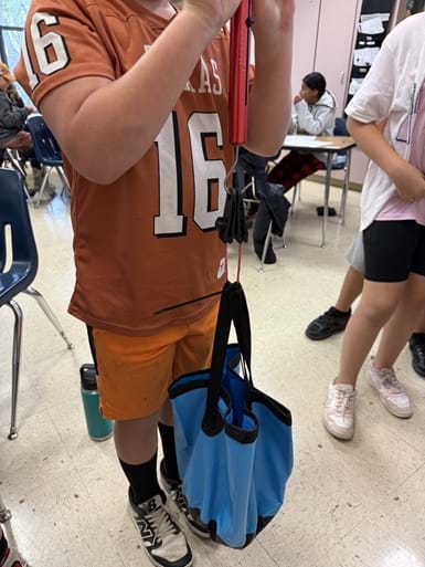 A photo showing a student holding a spring scale attached to a tissue paper prototype. The tissue paper is looped around a flexible bucket filled with weight to test the prototype. A photo showing a student holding a spring scale attached to a tissue paper prototype. The tissue paper is looped around a flexible bucket filled with weight to test the prototype.