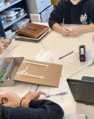 A photo showing a group of students working collaboratively at a white table. In the center, a piece of brown cardboard serves as a base for a structural prototype, with white plastic straws taped onto it in a geometric pattern. Surrounding the project are various supplies: a bundle of loose straws, a black tape dispenser, and a gray organizer bin containing scissors, a purple pen, and a laptop. A photo showing a group of students working collaboratively at a white table. In the center, a piece of brown cardboard serves as a base for a structural prototype, with white plastic straws taped onto it in a geometric pattern. Surrounding the project are various supplies: a bundle of loose straws, a black tape dispenser, and a gray organizer bin containing scissors, a purple pen, and a laptop.