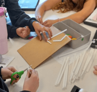 A photo showing students working collaboratively at a white table around a piece of brown cardboard. The cardboard has white plastic straws taped onto it. Surrounding the project are various supplies. A photo showing students working collaboratively at a white table around a piece of brown cardboard. The cardboard has white plastic straws taped onto it. Surrounding the project are various supplies.