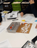 A photo showing students working collaboratively at a white table around a piece of brown cardboard. The cardboard has white plastic straws taped onto it. Surrounding the project are various supplies. A photo showing students working collaboratively at a white table around a piece of brown cardboard. The cardboard has white plastic straws taped onto it. Surrounding the project are various supplies.