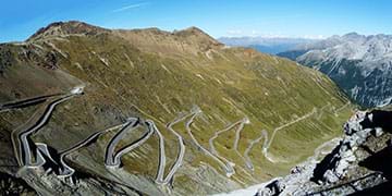 A photograph shows a winding road ascending through a valley and up a mountain. The road is made of several switchbacks that climb on top of each other, with large mountains in the background.  