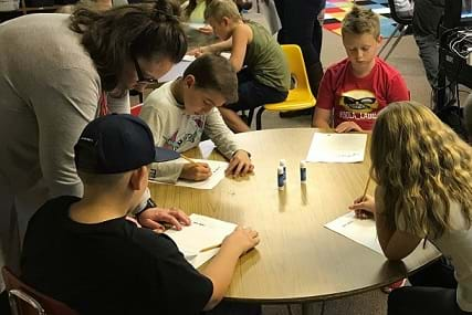 Students are sitting around a round table filling out worksheets as part of the activity.