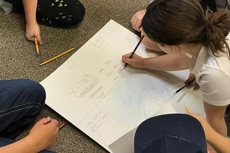 A small group of students sitting on a carpeted floor working on a poster that is focused on X-rays. One girl is using a pencil to write on the poster board.