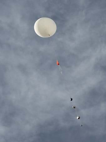 Looking up toward a dark blue sky with high white clouds at a white, round weather balloon that is carrying a long string of payload items below it, including a red parachute and four smaller boxes.