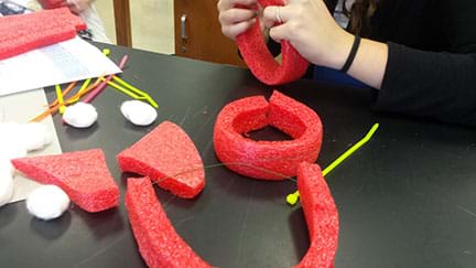 Photo of students building a better cast design in class, using dollar store materials – red foam, floral wire, cotton balls and pipe cleaners.