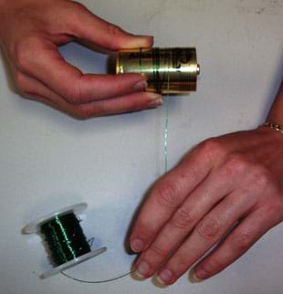 Photo shows a person's hands winding a coil of wire around a cylindrical battery. 