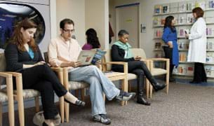 Photo shows four adults sitting and reading in chairs in a medical office waiting room, while in the background a woman in a lab coat talks to another person.