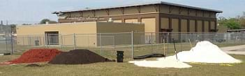 A photograph shows the yard outside a school with buckets, shovels and four piles of media: mulch (reddish fibers), topsoil (dark brown), limestone (pale beige), and sand (white). These materials were used to create a municipality-scale rain garden at Young Middle Magnet School in East Tampa, FL.