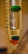 One red and two green liquid orbs suspended at different heights in a vial of pale yellow liquid.