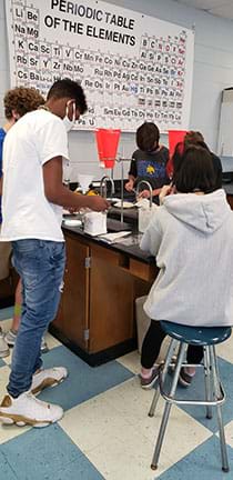 Students working together in a group. The students are sitting and standing around a lab bench while measuring ingredients.