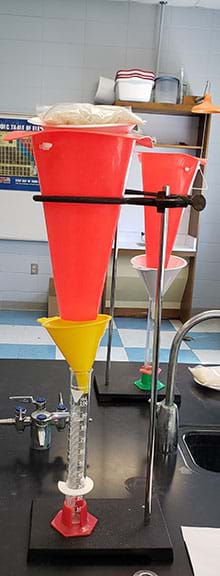 Complete setup of testing apparatus Option 1. A ring stand holds an inverted traffic cone with the top point cut off to form a large funnel. Below the cone is a small funnel which drains into a graduated cylinder. 
