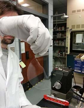 A photograph shows a man in a lab holding in one gloved hand a synthetic membrane synthesized at the University of Southern Mississippi. It looks like a clear plastic disk about 4 inches (~10 cm) wide.