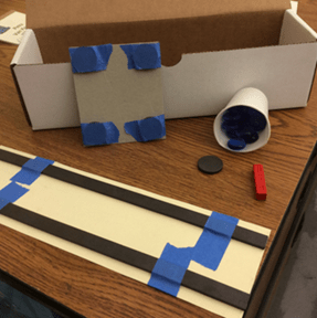 A photo showing a student-created maglev train model and maglev testing track. In the back of the photo, four disk magnets are taped to the corners of a square piece of cardboard. In the foreground, two flexible magnet strips are taped to a piece of tag board.