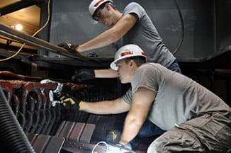 Two men in pants, t-shirts, hardhats and gloves use their hands to work on a large device; they are fixing a generator at a power plant.