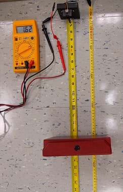 A photo shows a plastic handheld-sized multimeter device with a big dial and a small display screen, from which a black and a red wire are connected via alligator clips to a small black box (the infrared sensor). The black box is positioned at one end of a measuring tape laid flat on the ground facing a red box placed on the tape a foot or so away.