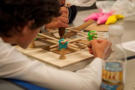 A photograph shows two students at a table focused on using a model/mock-3D bioprinter. One student squeezes an icing bag that represents the extrusion of cells, while the other student moves the cardboard printing stage in order to print an object of the desired shape.