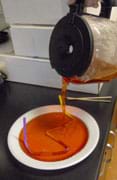 Photo shows boiling red liquid being poured into a shallow plate, submerging a multi-angled straw taped to its base.
