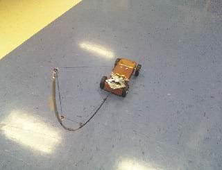 A small four-wheeled energy storage car on linoleum floor. Looks like the car has a curved mast made from a fishing pole with wire attaching it to the car.