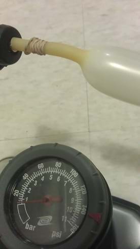 A photograph shows a latex tube attached to a bicycle pump's needle tip extension and the pump's pressure gauge nearby for easy visibility to observe the amount of pressure when the tube explodes.