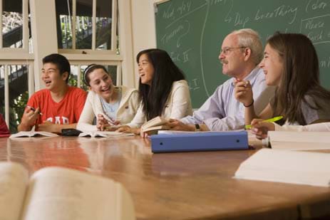 Photo shows four students and a teacher sitting and laughing around a table with books open and a chalkboard backdrop. A Harkness table being used at The College Preparatory School.