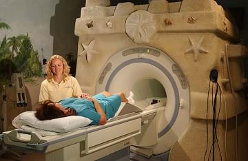 A photograph shows a female patient laying on a table getting ready to have an MRI as a medical support staff stands nearby.