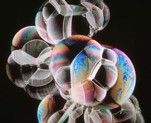 A close-up photograph of a soap bubble on a black background. Swirls of rainbow colors are visible on many conjoined domed surfaces.