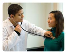 A photograph shows a man in a white lab coat holding a stethoscope to the chest of a female patient to listen to her heart.