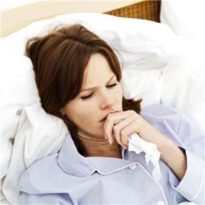 Photo shows a woman lying sick in a bed with a tissue in her hand.