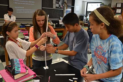 Four students are constructing a paper tower that is narrow, round, and about two feet tall.