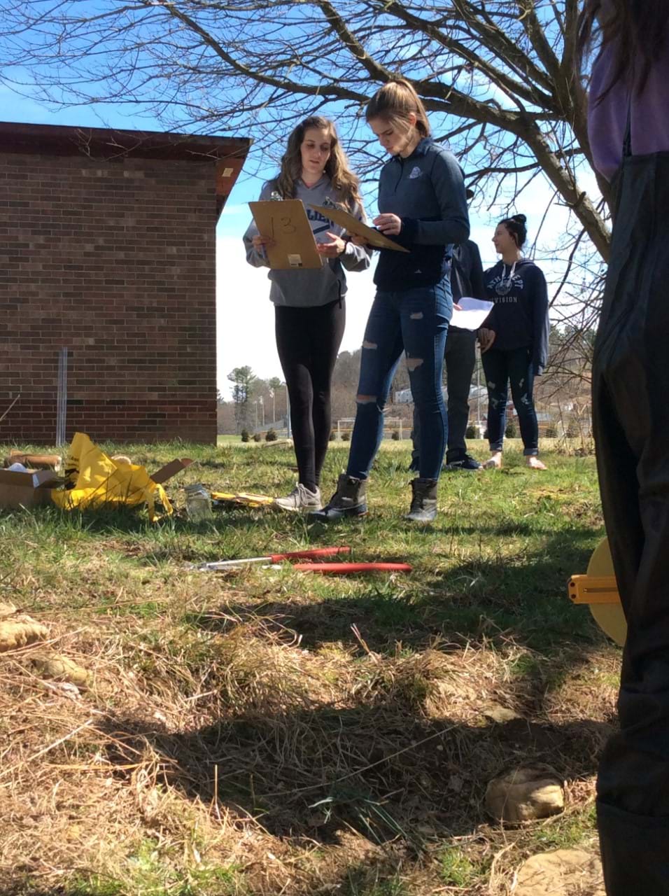 A group of students stands in a school yard as they inventory measuring tools that lay on the grass.