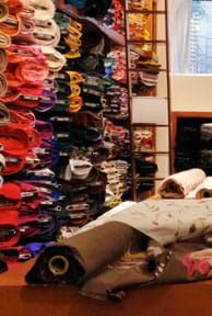 Photograph shows the inside of a fabric store, with bolts of fabric on a foreground table and stacked in shelves to the ceiling.