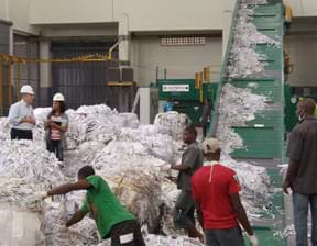 A photograph shows six people moving piles of shredded paper that arrive in a room via conveyor belt.