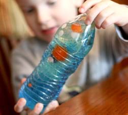 A photograph shows a boy examining the contents of a clear plastic water bottle that he holds with both hands. The bottle contains colored water, vegetable oil and little toy trinkets.