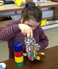 A photograph shows a child playing with a toy made from a clear plastic bottle that is partially covered with colored tape and contains bells and small balls.