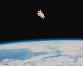 Photo shows a person in a spacesuit floating in the black atmosphere above the curve of the planet Earth below.