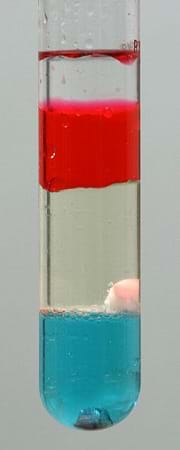 A test tube shows colored layers. From top: clear/transparent, red, slightly yellow, a white blob, light blue, a metallic chunk.