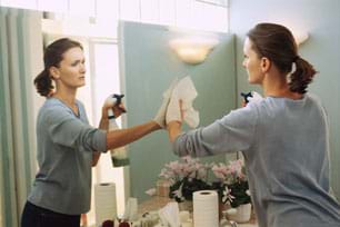 Photo shows a woman using paper towels and a spray bottle of liquid to clean a wall mirror. Photo shows a woman using paper towels and a spray bottle of liquid to clean a wall mirror.